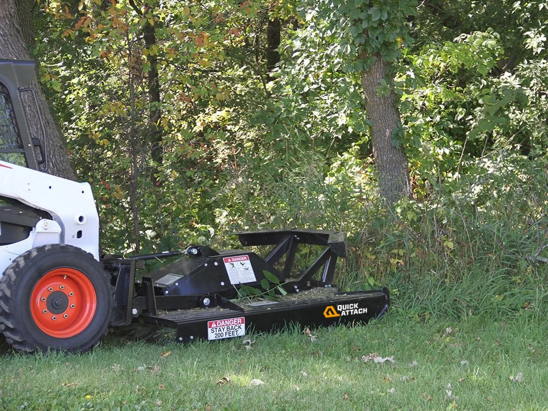 Rotary brush cutter deck on a skid steer pushing into tall weeds at the edge of the woods, bush hogging