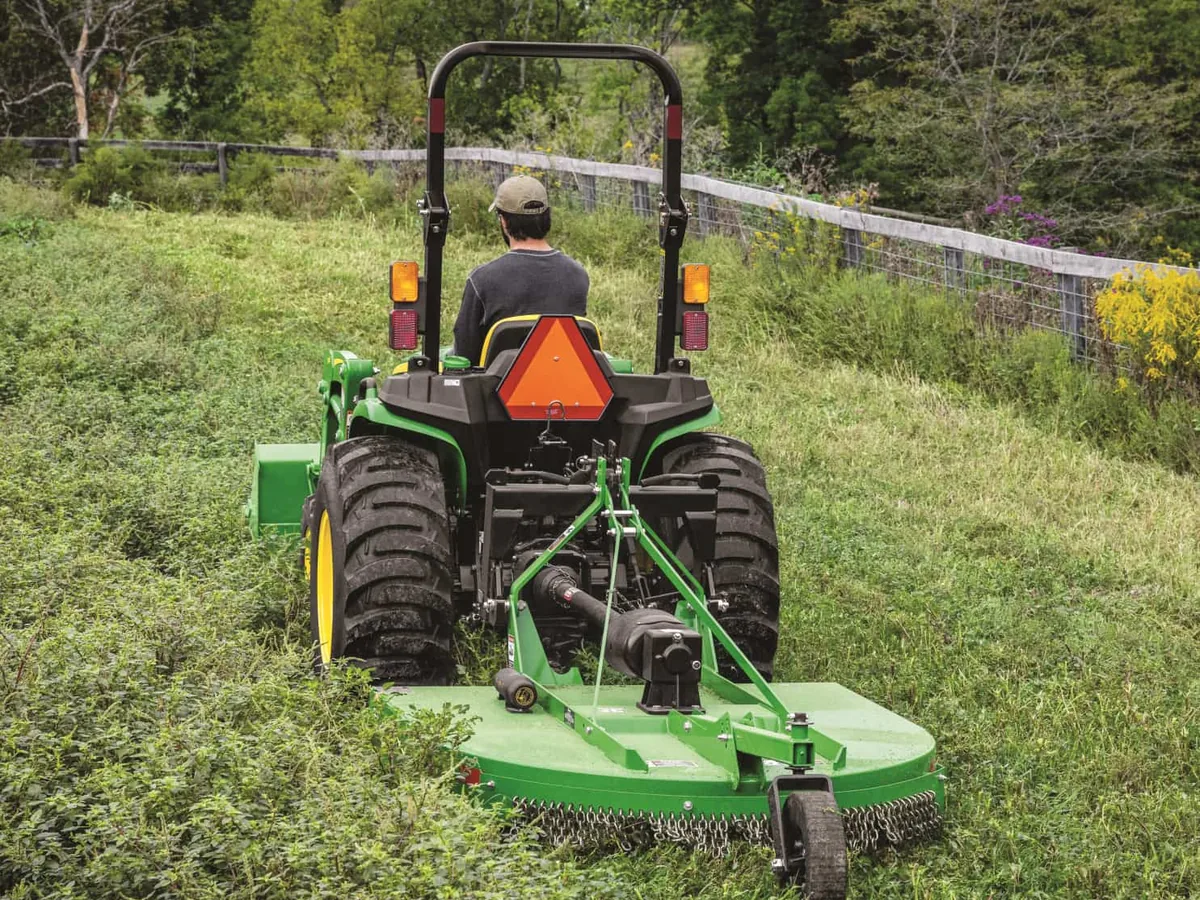 Tractor pulling a rotary cutter through tall weeds along a board fence, bush hogging