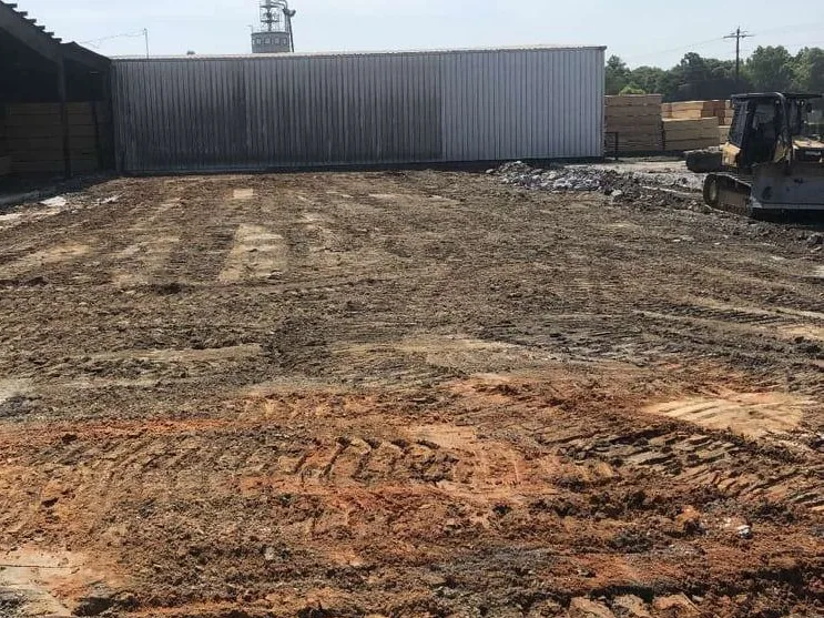 Graded dirt yard in a lumber mill beside a long metal building and stacked lumber, civil construction