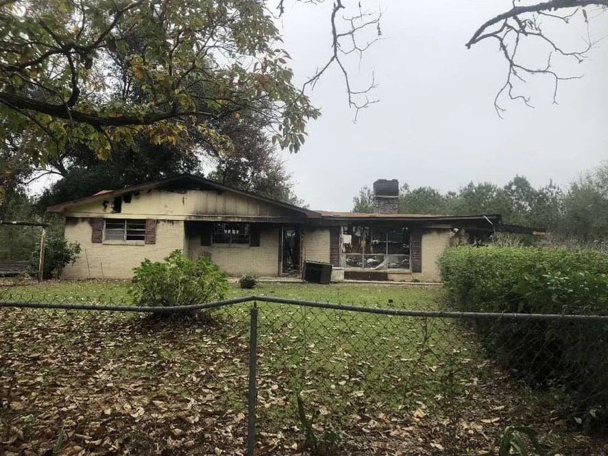 Fire-damaged brick house behind a chain-link fence before demolition
