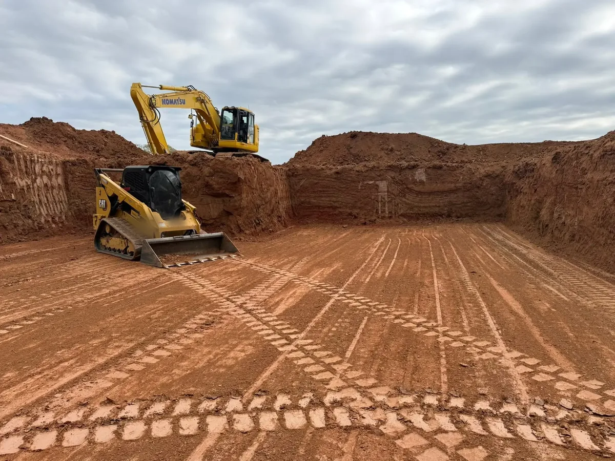 Deep cut in red clay with an excavator on the rim and a track loader grading the floor, excavation