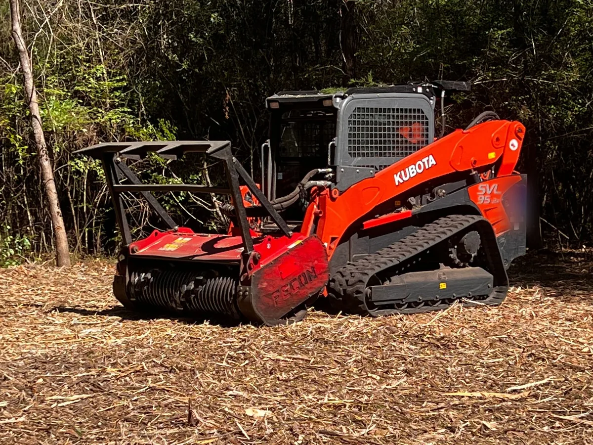 Compact track loader with a mulching head parked on a fresh layer of mulch at the edge of a thicket, forestry mulching