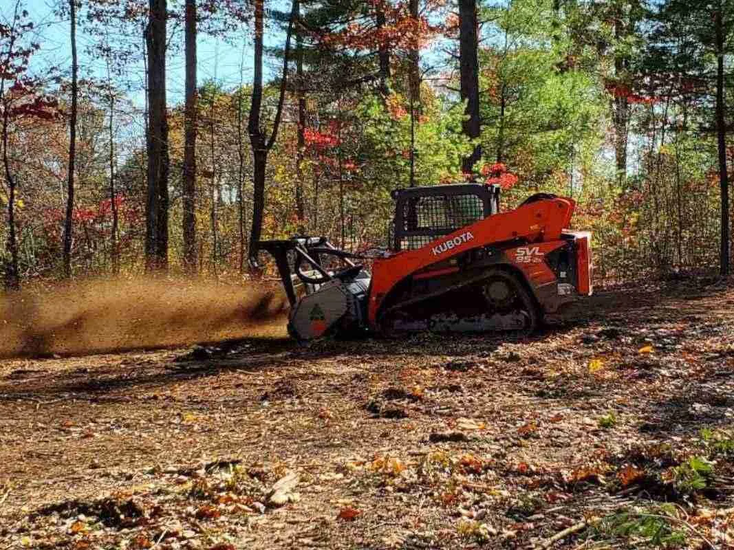 Compact track loader with a mulching head throwing wood chips in autumn woods, forestry mulching
