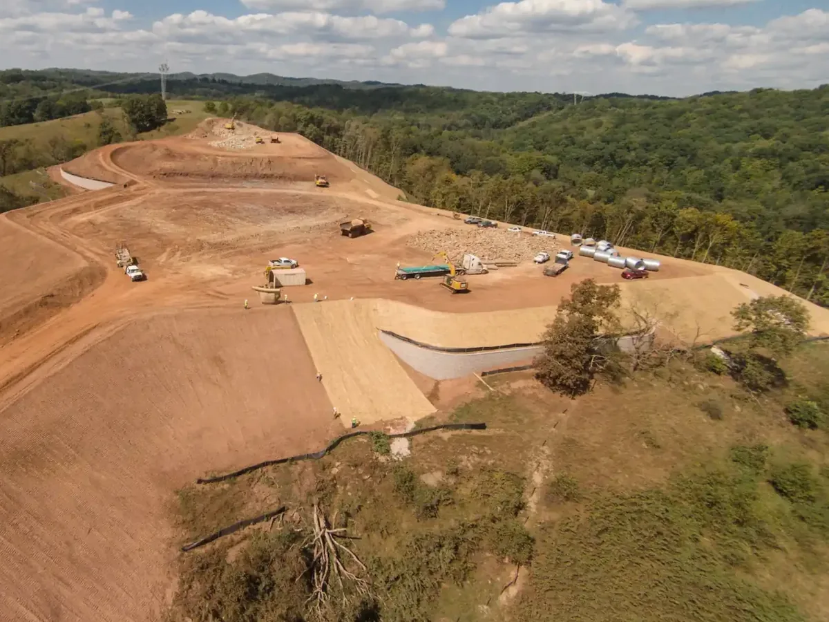 Aerial view of a well pad being cut into a wooded ridge with excavators and dump trucks, oilfield services