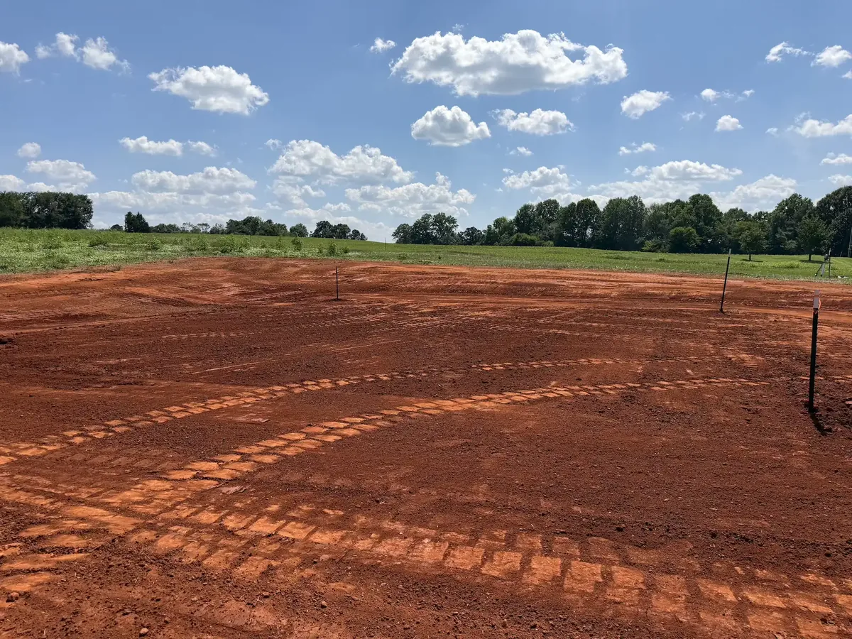 Graded red clay building pad with track marks and grade stakes beside a pasture, site preparation