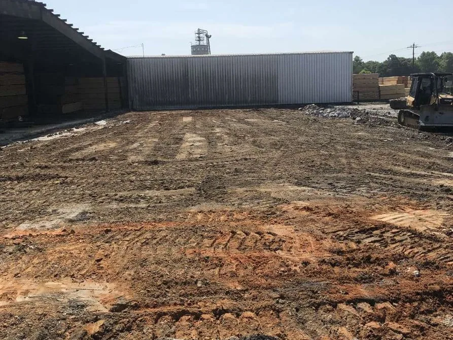 Freshly graded dirt pad with track marks beside a metal building and lumber stacks, site preparation