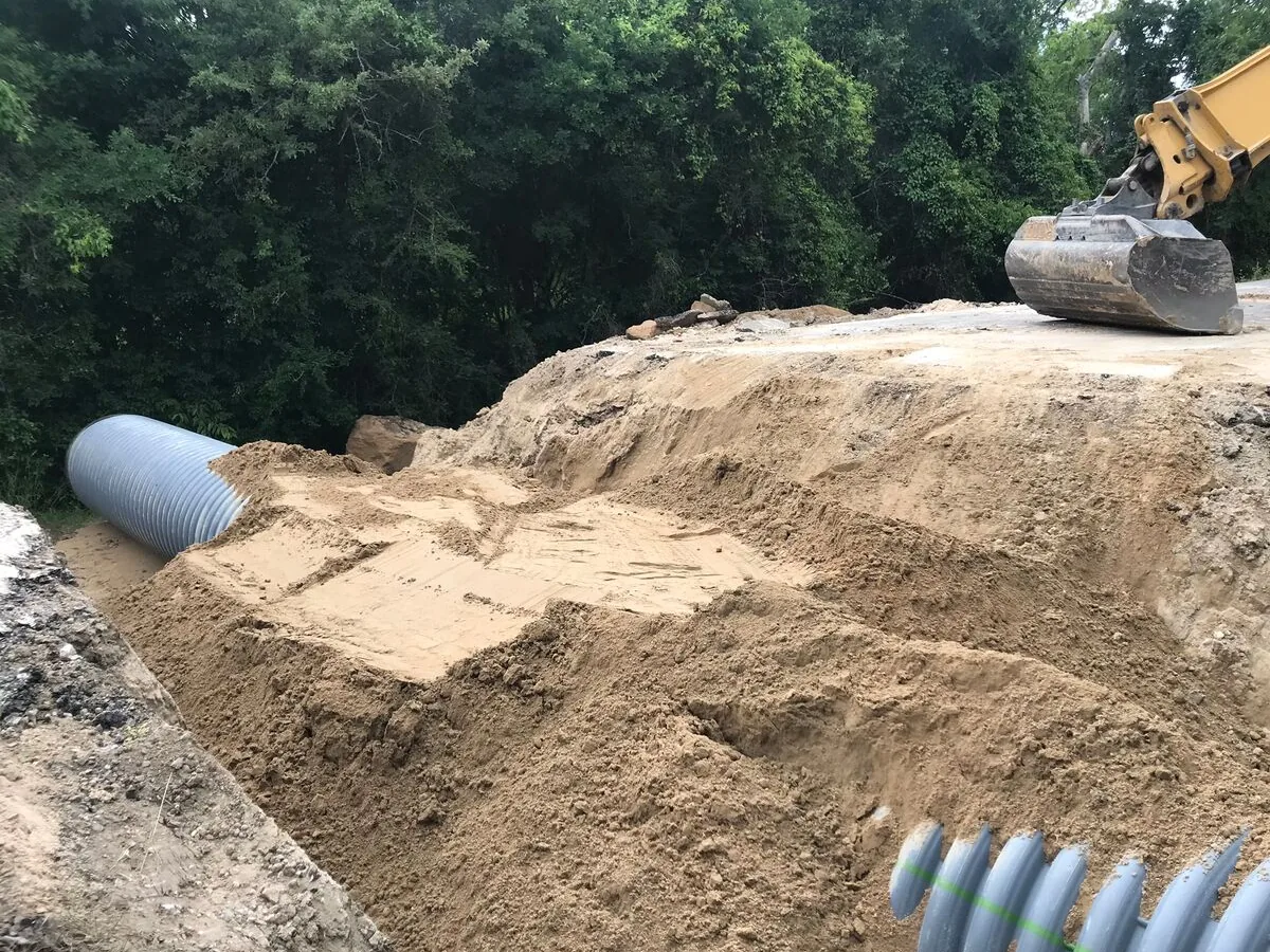 Culvert pipe set in a trench under fresh sand backfill with an excavator bucket working, underground utilities