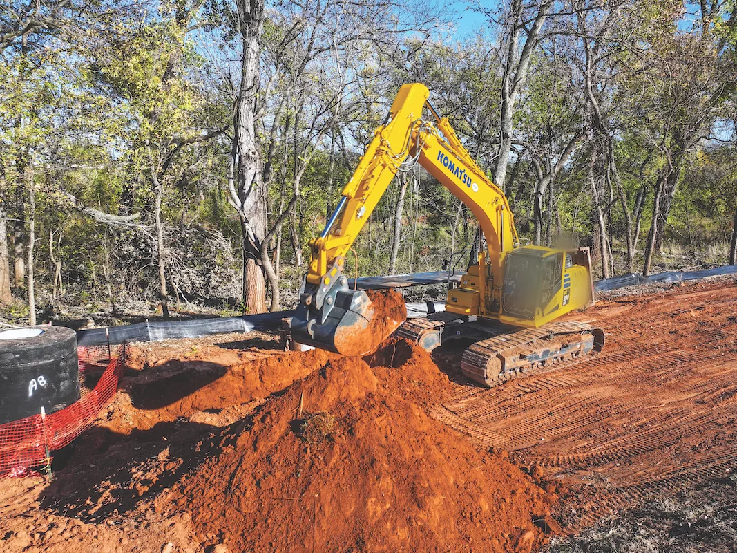Excavator digging a utility trench in red clay beside a utility riser and silt fence, underground utilities